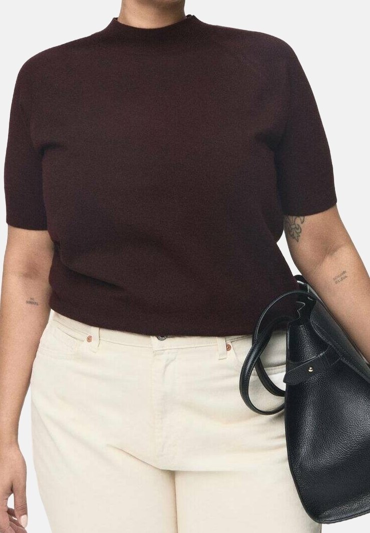 Personne portant un haut sombre à manches courtes et un pantalon clair, tenant un sac à main en cuir noir sur son côté droit, tatouages visibles sur les bras.