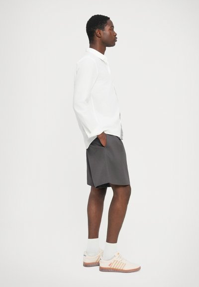 Chemise blanche à manches longues, short gris et baskets crème avec un accent orange. Coupe droite, design décontracté et poches visibles sur le short.