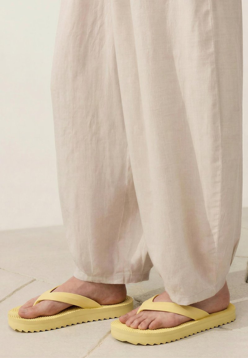 Pieds portant un pantalon beige ample et des tongs jaunes avec des semelles épaisses et texturées, debout sur un sol carrelé.