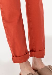 Orange cotton pants with rolled cuffs. Cuffs feature a subtle paisley pattern. Soft fabric texture, relaxed fit, paired with beige slip-on shoes.