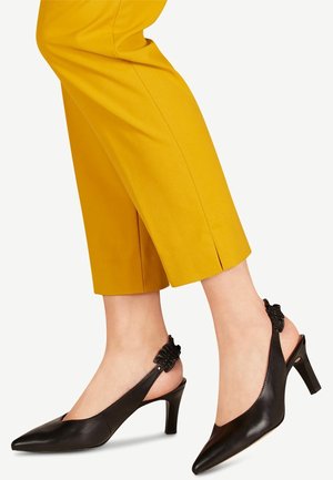 Czarne, spiczaste sandały na obcasie z zakrzywionym obcasem i teksturowanymi czarnymi akcentami z tyłu, noszone z żółtymi spodniami o krótkim kroju.
