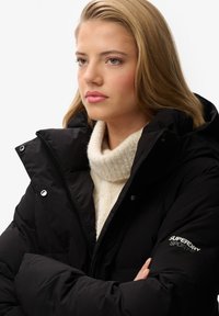 Chaqueta de plumón negra con un alto cuello, que presenta una cremallera y botones a presión. Llevada sobre un suéter de punto de color crema. Acentuada con un logo.