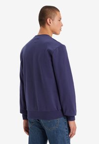 Paarse sweatshirt met lange mouwen, geribbelde boorden en onderzoom. Zachte textuur met een ronde hals. Draagt met blauwe spijkerbroek.