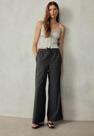 Young woman with long brown hair standing against a light gray wall, wearing a gray tank top, dark wide-leg pants, and black sandals.