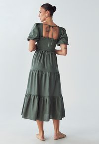 Maslinzasto zelena, višeslojna maksi haljina s naboranom korzetom i puf rukavima, s prorezom na leđima koji se učvršćuje vezicom.