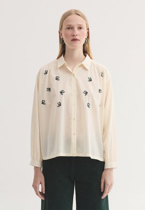 Femme portant une chemise crème à boutons avec de petits motifs brodés verts et un pantalon vert foncé, debout devant un fond uni.