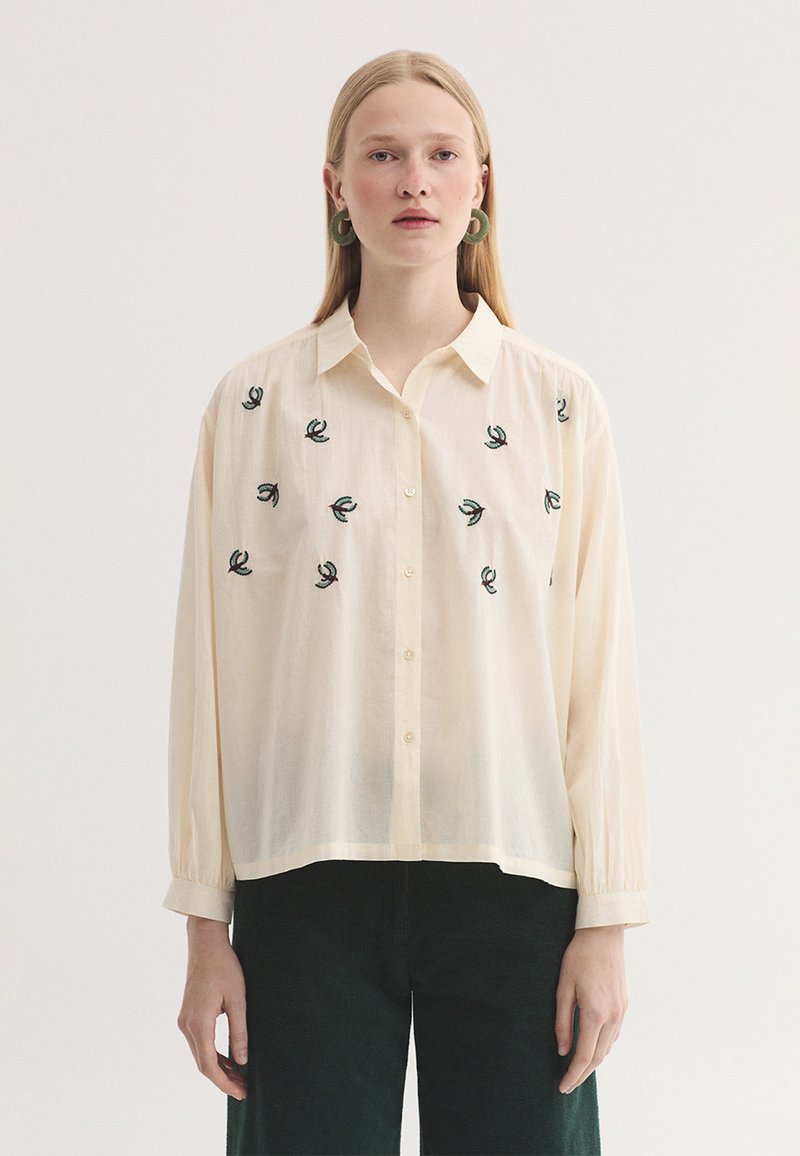 Femme portant une chemise crème à boutons avec de petits motifs brodés verts et un pantalon vert foncé, debout devant un fond uni.