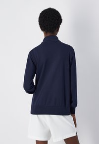 Sudadera de manga larga azul marino con cuello tipo mock. Material suave de mezcla de algodón, corte relajado, puños y cintura acanalados. Combinada con pantalones cortos blancos.