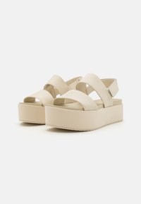 Beige platform sandals with wide cross straps and a cushioned footbed. Smooth texture, rubber sole, and elevated heel design.