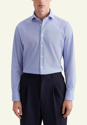 Man wearing a light blue button-up dress shirt tucked into high-waisted black pleated trousers, standing against a plain background.