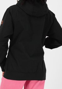 Persona che indossa una giacca nera con cappuccio con una toppa circolare marrone sulla manica e pantaloni rosa, vista da dietro.