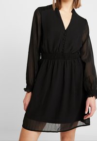 Schwarzes knielanges Kleid mit durchsichtigen langen Ärmeln, gerafftem Taillenbereich, Knopfleiste und subtilen vertikalen Streifen auf dem Stoff.