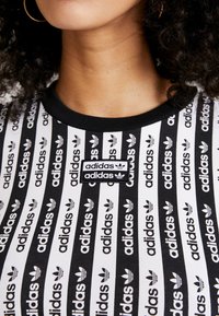 Detailní záběr na osobu v černo-bílém pruhovaném tričku Adidas s opakujícími se logy Adidas a dvojitým logem na hrudi.