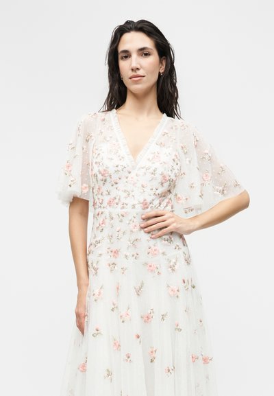 Γυναίκα με σκούρα μαλλιά φοράει λευκό φόρεμα με V-λαιμόκοψη, διαφανή puff μανίκια και ροζ λουλουδένια κεντήματα, ποζάροντας με το χέρι στη μέση.