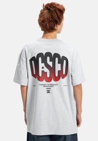 Světlé šedé oversized tričko s velkým červeným a černým potiskem na zádech, obsahující logo "DC" a text. Krátké rukávy, bavlněný materiál.