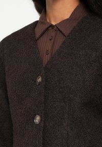 Close-up van een persoon in een donkerbruin, gestructureerd vest over een geribbeld, met knopen gesloten shirt met een puntige kraag.