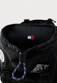 Rucsac negru cu închidere cu șnur, având un accesoriu de frânghie albastră și roșie și o etichetă cu logo-ul "TOMMY JEANS" în interior. Materialul este neted și rezistent.