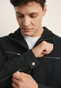 Czarna kurtka zapinana na zamek z wysokim kołnierzem, z metalowymi elementami w kolorze srebrnym i kieszeniami zapinanymi na zamek. Materiał wydaje się gładki o matowym wykończeniu.