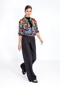 Giacca bomber floreale con ricami vivaci in rosso, blu e verde; top nero aderente; pantaloni neri larghi; sandali flat.