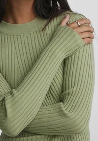 Personne portant un pull côtelé vert clair à manches longues, les bras croisés, montrant une bague argentée ondulée sur le majeur de la main droite.