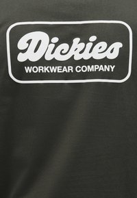 Σκούρο ύφασμα με λευκό έντονο κείμενο «Dickies» πάνω από μικρότερο κείμενο «WORKWEAR COMPANY» μέσα σε στρογγυλεμένο ορθογώνιο περίγραμμα.