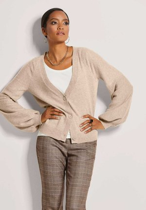 Beige gebreid vest met een diepe V-hals, vier knopen en brede mouwen. Draagt over een wit topje en gecombineerd met een bruine ruitbroek.