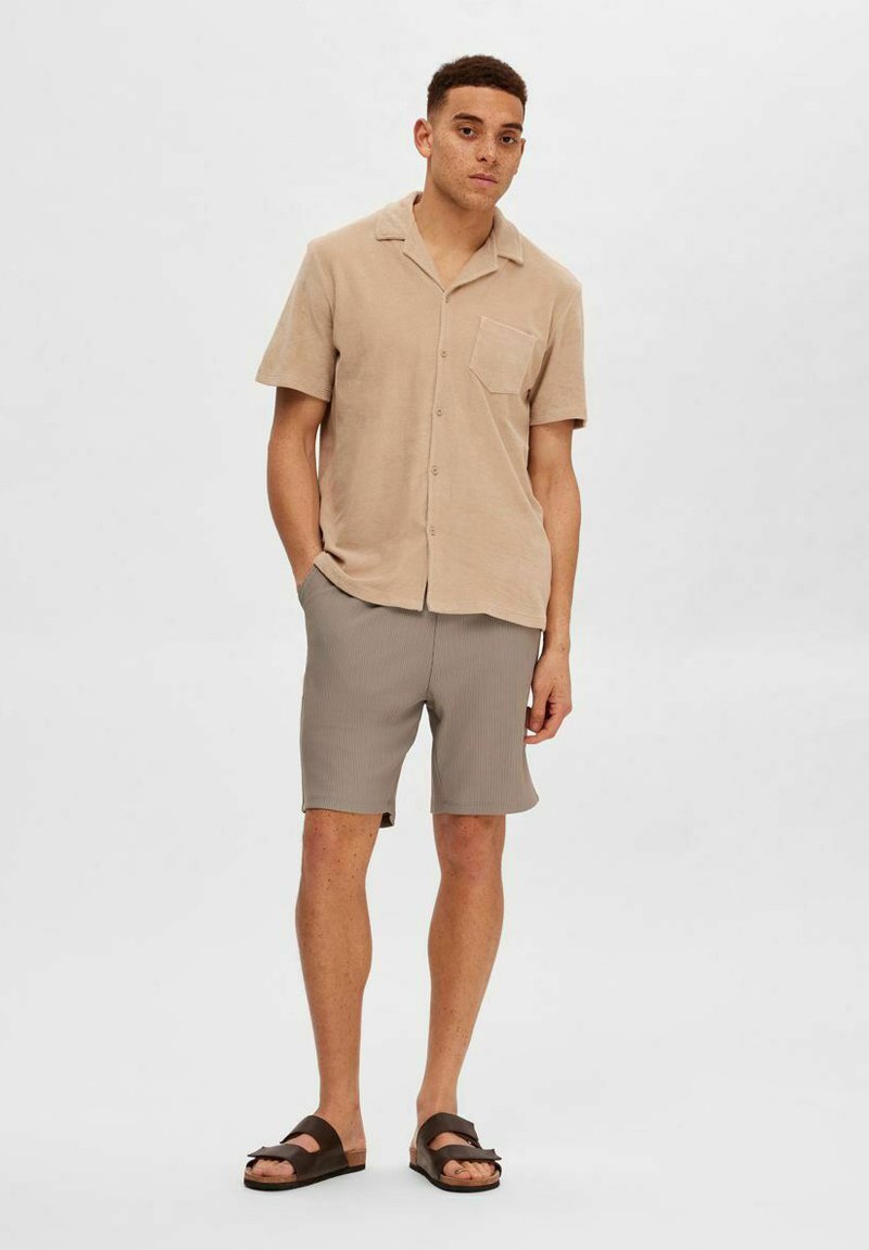 Chemise beige à manches courtes avec une poche, accompagnée de shorts en ribbed gris et de sandales marron. Tissu doux et coupe décontractée.