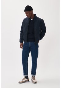 Jūros pajėgų bombonešio striukė, tamsi megztinis, mėlyni džinsai ir kreminiai sportbačiai. Modelis stovi profiliu, demonstruodamas atlenktą kelnių apačią ir tekstūrinę skrybėlę.