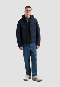Marineblauer Puffer-Jacke mit Kapuze, ausgestattet mit einem gesteppten Design und einer Fronttasche. Kombiniert mit einem olivgrünen Hemd und hellblauen Jeans.