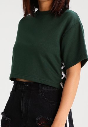 Osoba nosząca ciemnozielony krótki T-shirt z białymi sznurowanymi bokami oraz wysokim stanem czarne jeansy z przetartymi detalami na jednym kolanie.
