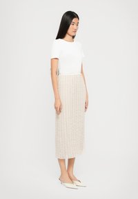 Jupe midi beige avec un motif texturé vertical, associée à un T-shirt blanc ajusté et des chaussures blanches à enfiler. Tissu lisse avec des détails subtils.