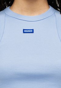 Ανοικτό μπλε ριμπ τοπάκι με ένα μικρό ορθογώνιο μπλε και λευκό λογότυπο «HUGO» στο στήθος. Το ύφασμα φαίνεται υφασμένο και εφαρμοστό.