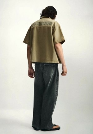 Person mit lockigem Haar, die ein beigefarbenes kurzärmeliges Hemd mit Text auf dem Rücken, weite dunkle Jeans und schwarze Sandalen trägt.
