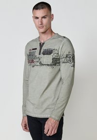 Hosszú ujjú, szürke póló gombos nyakkivágással, mellzsebbel és fekete-piros nyomott grafikával. Egyszerű, texturált anyag.