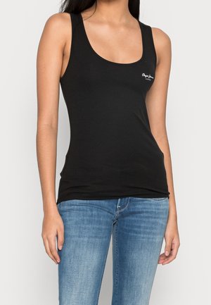 Kvinde iført en tætsiddende sort tanktop med lille hvidt logo på brystet, kombineret med lyseblå cowboybukser, stående mod en ensfarvet baggrund.