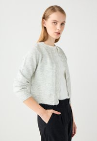 Hellgrauer gestrickter Cardigan mit rundem Ausschnitt, gewellter Knopfleiste und gerippten Bündchen. Getragen über einem weißen Oberteil und schwarzen Hosen.