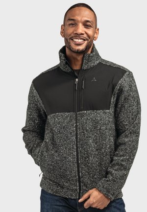 Lächelnder Mann mit einer grauen und schwarzen Fleecejacke mit Reißverschluss, die Hände in den Taschen, steht vor einem einfarbigen Hintergrund.