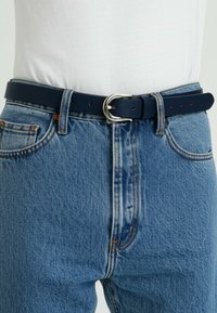 Marineblauer Leder-Gürtel mit einer silbernen Metallschnalle, getragen über hellblauen Jeans aus Denim, die einen strukturierten Stoff und ein klassisches Fünf-Taschen-Design aufweisen.