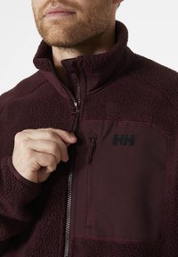 Fleece'owa kurtka w ciemnym burgundzie z zamkiem z przodu, wysokim kołnierzem i kieszenią na piersi z czarnym logo. Miękka tekstura z kontrastującymi materiałami.