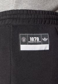 Černé tepláky s texturovaným povrchem, s tkanou etiketou zobrazující "1878" a "Manchester United" vedle loga Adidas.