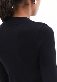 Top nero a maniche lunghe con scollatura rotonda, caratterizzato da una texture liscia e aderente con cuciture minime, mostrato parzialmente da dietro.