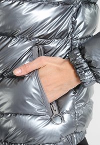 Metallic grå puffjakke med quiltede paneler, en lynlåslomme og elastiсkuffer. Glat, skinnende materiale med en blank finish.