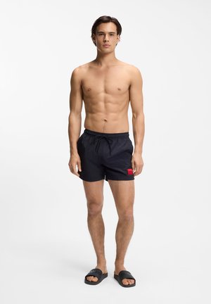 Hemdloser Mann, der mit dem Gesicht nach vorne steht, trägt schwarze Badeshorts mit rotem Logo-Patch und schwarze Slide-Sandalen vor weißem Hintergrund.