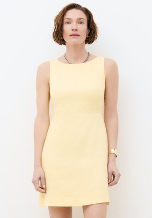 Femme aux cheveux courts bruns portant une robe sans manches jaune pâle, un collier en chaîne dorée, des boucles d'oreilles créoles et un bracelet en or, debout contre un mur blanc.