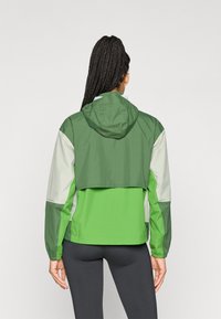 Chaqueta ligera verde y gris con capucha, que presenta un diseño corto, puños elásticos y una textura suave.