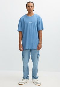 Camiseta de algodón de color azul claro con mangas cortas y "SERVICIO 24 HORAS" impreso en blanco. Combinada con pantalones cargo azul claro y zapatillas blancas.