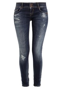 Jeans ajustados en denim azul oscuro con zonas desgastadas y parches dañados. Presenta cinco bolsillos, cierre de botón y cremallera.