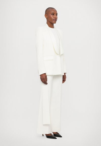 Witte op maat gemaakte blazer met een sjawlzoom, gecombineerd met bijpassende wijde broek en zwarte puntige hakken. Gladde textuur en minimalistische afwerking.