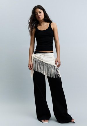 Jeune femme aux longs cheveux portant un haut noir sans manches, un pantalon noir à jambes larges, une ceinture blanche à franges et des chaussures blanches à bouts ouverts.
