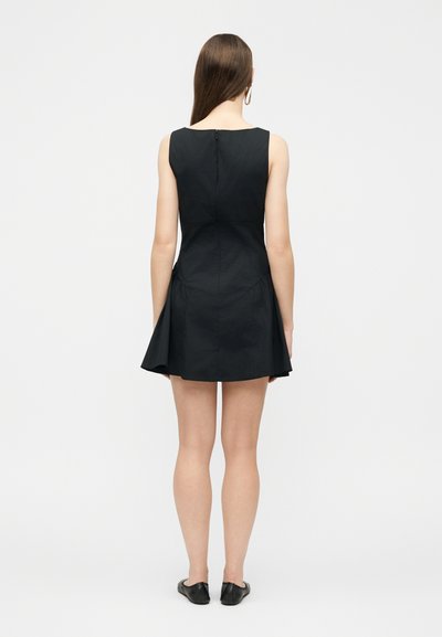 Γυναίκα με μακρυά καστανά μαλλιά, φορώντας μαύρο αμάνικο φόρεμα και μαύρα επίπεδα παπούτσια, στάση με την πλάτη προς το μέρος μας σε λευκό απλό φόντο.